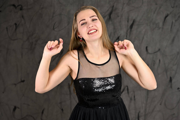 A photo of a pretty smiling girl with long hair and excellent make-up in a black dress stands in different poses. Universal concept of horizontal female portrait on gray alternative background. - Valokuva, kuva