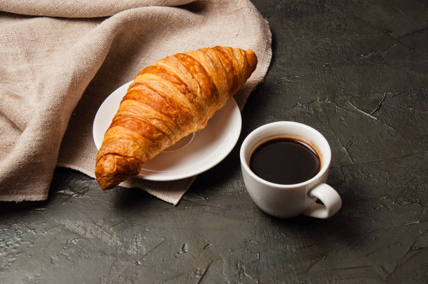 Croissant en kopje koffie met lepel en schotel op een donkere achtergrond met grijs linnen doek - Foto, afbeelding