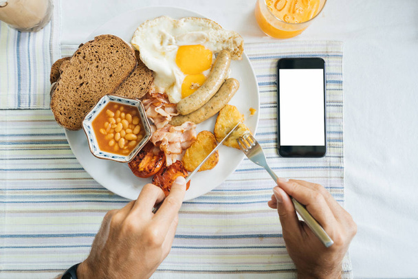 man has breakfast, hands with a knife and a fork, a dish and a smartphone with a blank screen. Calorie  - Photo, image