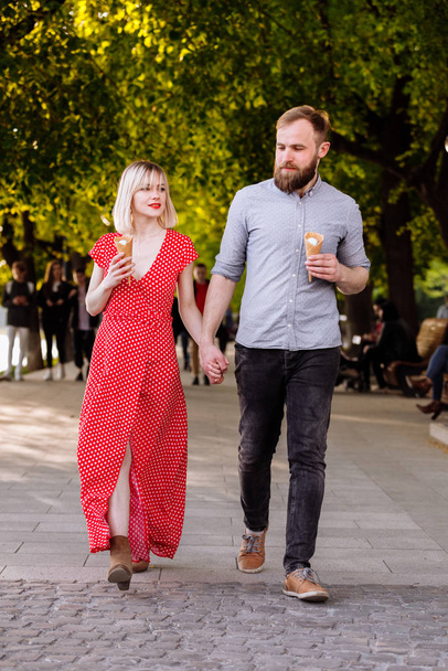 smiling hipsters couple eating ice cream and having fun in the city. stylish young man with beard and blonde woman in red dress are walking in the street. - Foto, imagen