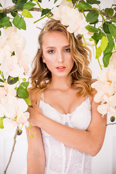 Attractive young girl with blond curly hair and beautiful make-up in a tender light lace corset with white orchids - Foto, afbeelding