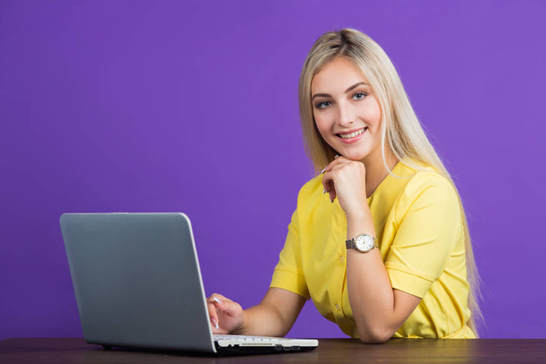 beautiful young woman in a yellow dress on a purple background with her hair sits at a table with a laptop - Photo, image