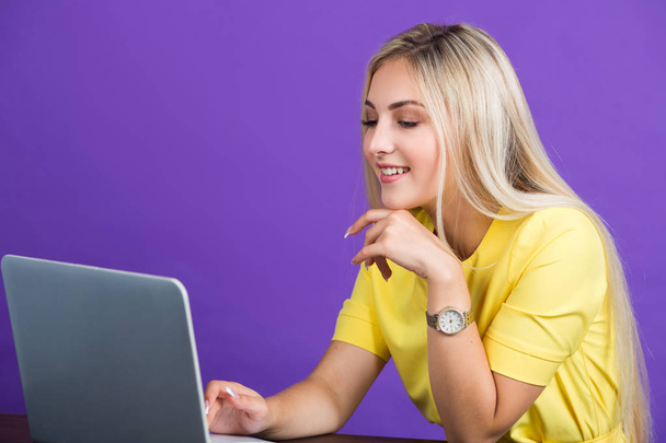 beautiful young woman in a yellow dress on a purple background with her hair sits at a table with a laptop - Zdjęcie, obraz