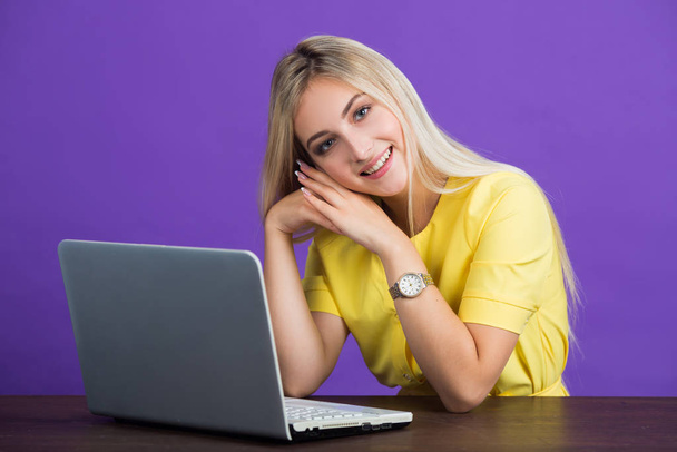 beautiful young woman in a yellow dress on a purple background with her hair sits at a table with a laptop - Photo, Image