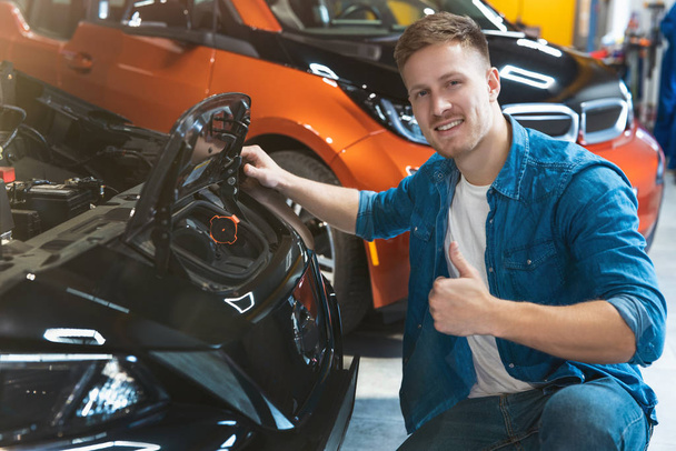young positive man customer sitting near vehicle open bonnet exploring electric motor in car dealership center showing like sign, zero emission concept - Zdjęcie, obraz