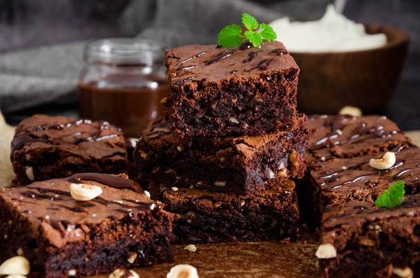 Házi készítésű csokis brownie desszert mogyoróval, csokoládé mázzal és tejszínhabbal sütőpapíron sötét háttérrel. Első látásra. Vízszintes tájolás. - Fotó, kép
