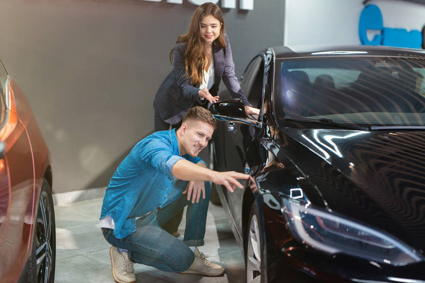 young positive woman manager in business suit consulting young man client showing vehicle exterior in electric car dealership center, professional cooperation - Фото, зображення