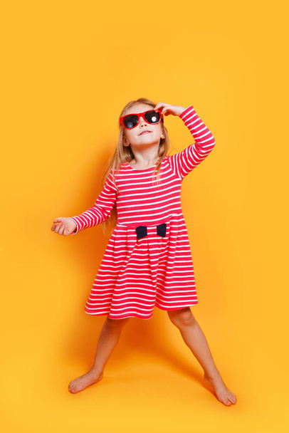 funny child girl wearing sunglasses and red dress on a orange background - Fotoğraf, Görsel