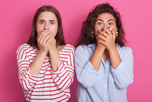 Horizontal indoor shot of surprised mysterious good looking young women covering their mouths, being scared, keeping secrets, being shocked by news, opening eyes widely. People and secrets concept. - Fotografie, Obrázek