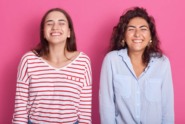 Photo of two cheerful energetic emotional ladies with black hair closing eyes, feeling good, being in high spirits, wearing casual clothes, smiling sincerely, being full of energy. Emotions concept. - Foto, Imagen