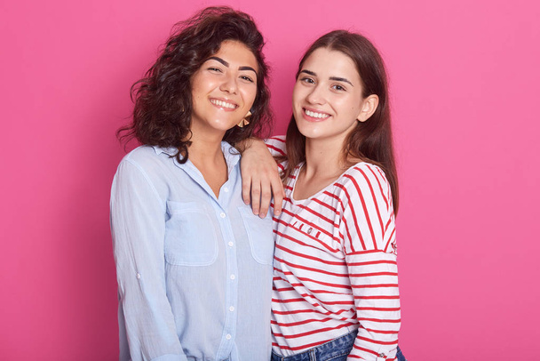 Indoor shot of two cheerful good looking energetic friends, smiling sincerely, hugging each other, looking directly at camera, wearing casual clothes, spending free time together. Friendship concept. - Fotoğraf, Görsel