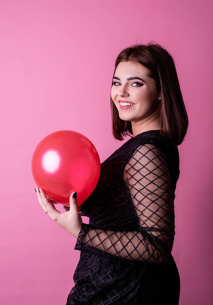 Sexy woman in black dress holding a red balloon isolated on pink background - Photo, image