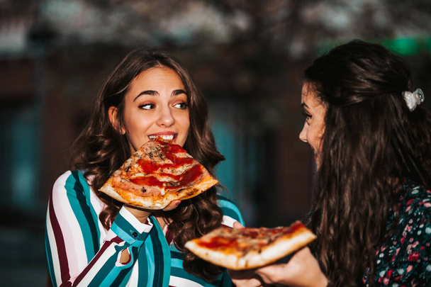 Portrait of two young women talking and eating pizza outdoors, h - 写真・画像