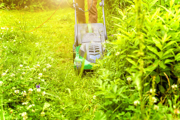 Człowiek przycinający zieloną trawę kosiarką na podwórku. Ogrodnictwo kraju tle stylu życia. Piękny widok na świeżą zieloną trawnik w świetle słonecznym, krajobraz ogrodowy w sezonie wiosennym lub letnim. - Zdjęcie, obraz