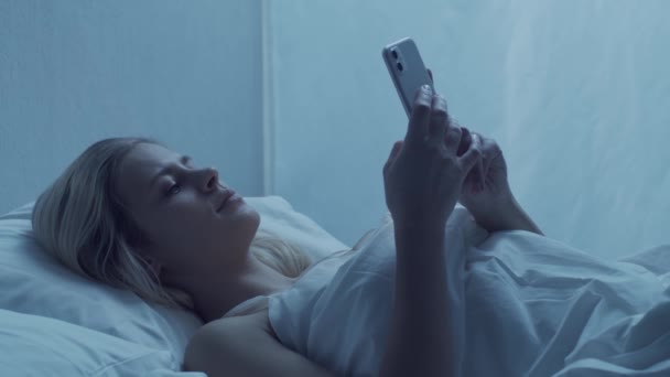 Young woman lying in the bed with a smartphone at night. Beautiful blond resting girl. Twilight in the bedroom, moonlight from the window. Health and rest concept. - Séquence, vidéo