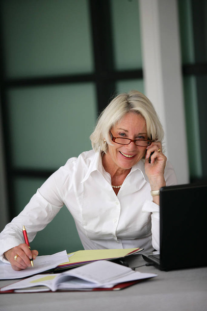 Portrait of a senior woman sitting at a desk phone with cell phone - Foto, afbeelding