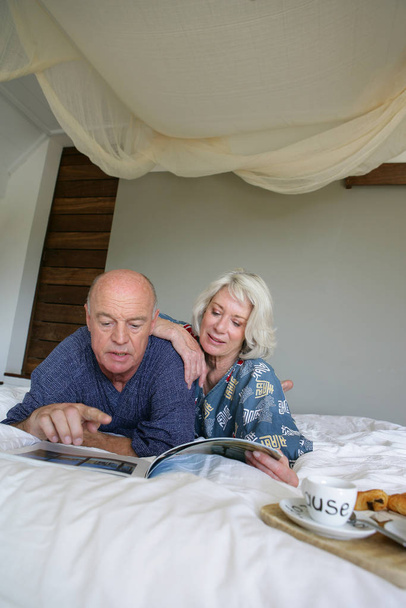 Older man and woman in bathrobe lying on a bed while leafing through a magazine - Fotoğraf, Görsel