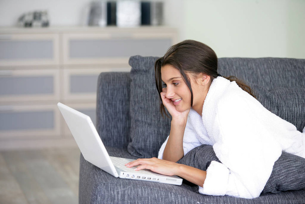 Bornozlu gülümseyen bir kadının portresi bir dizüstü bilgisayarın önünde kanepede yatıyor. - Fotoğraf, Görsel