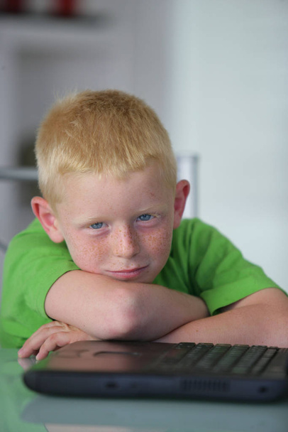 Portrait of a smiling little boy sitting at a desk near a laptop - Photo, Image