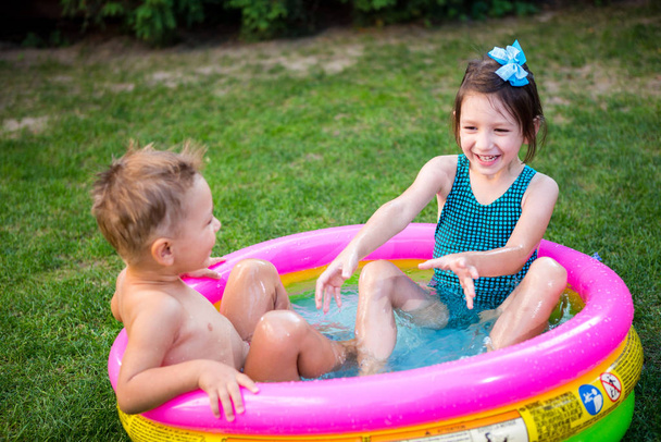 Két kistesó játszik és fröcsköl a medencében a forró nyári napon. Gyerekek úszkálnak a medencében. Két vidám aranyos gyerek játszik és szórakozik, fröcskölnek a felfújható medencében a kertben - Fotó, kép