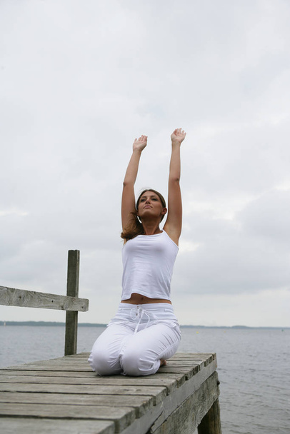 Woman meditating by lifting the arms on a dock at the waterfront - Foto, Bild