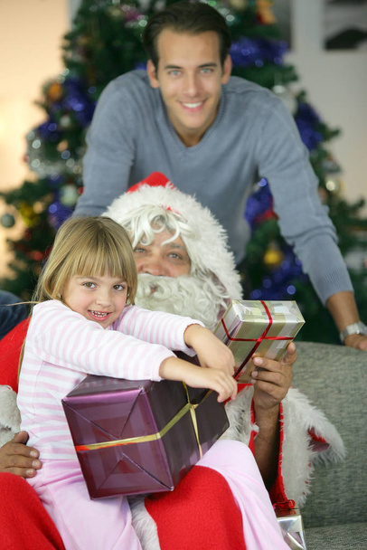 Portrait of a smiling girl with gifts sitting on Santa's lap - Фото, изображение