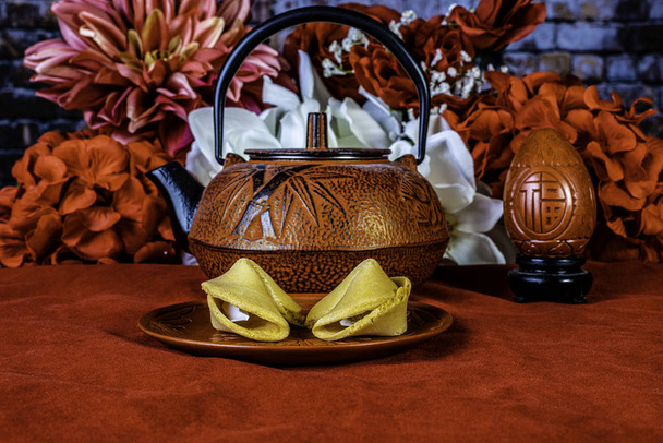 dvě čínské sušenky štěstí na červeném talíři s hrncem červeného čaje a ubrusem s červenými a bílými květy v pozadí - Fotografie, Obrázek