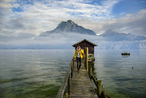 Άνδρας με κίτρινο σακάκι που περπατά σε ξύλινη πεζογέφυρα στη λίμνη Traunsee με κορυφή Traunstein σε φόντο που καλύπτεται με χαμηλά σύννεφα, που βρίσκεται στην Αυστρία, περιοχή των Άλπεων. - Φωτογραφία, εικόνα