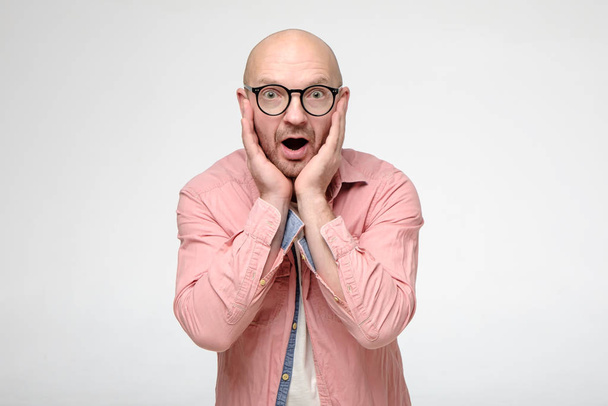 Shocked bald man in glasses emotionally holds hands on his face, in surprise opens mouth and looks into the camera with big eyes. - Photo, Image