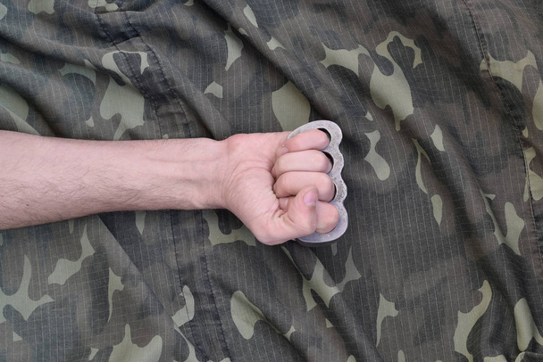 Punho masculino com juntas de latão no fundo de uma jaqueta de camuflagem. O conceito de cultura skinhead, armas corpo a corpo feitas à mão
 - Foto, Imagem