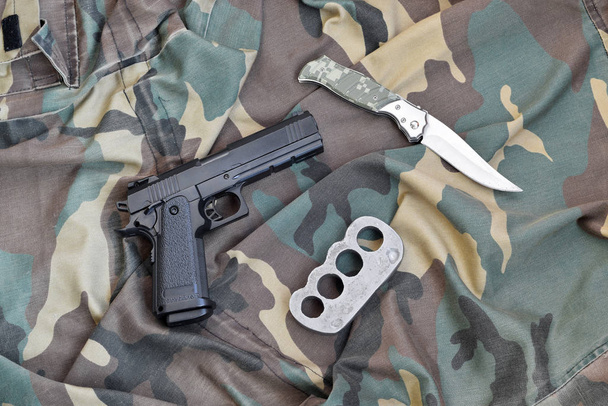 Pistola giace con nocche in ottone e coltello su camuffamento uniforme militare da vicino. Concetto di saccheggio e commercio di armi
 - Foto, immagini