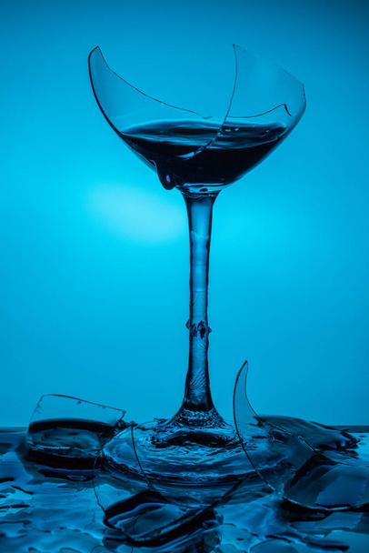 Broken glass with wine stands on a glass mirror table on a blue background - Фото, изображение