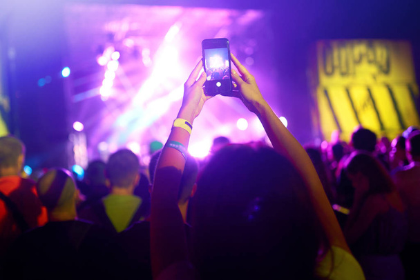 Hand with a phone records live music festival. People taking photographs with smart phone during a public music concert. Crowd raising their hands, dancing and enjoying great the concert. - Photo, Image