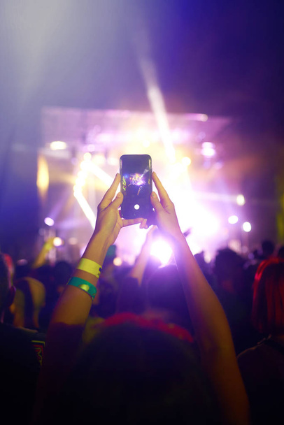 Mano con un teléfono graba el festival de música en vivo. Gente tomando fotografías con un teléfono inteligente durante un concierto de música pública. Multitud levantando las manos, bailando y disfrutando mucho del concierto
. - Foto, imagen