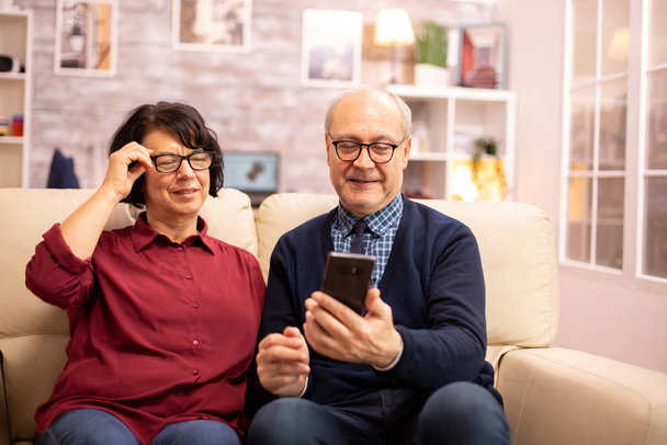 Piękna starsza para robi sobie selfie siedząc na kanapie - Zdjęcie, obraz