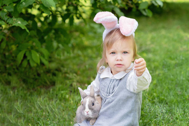 kleines Mädchen im Hasenkostüm, das mit Kaninchen spielt, während es im Garten auf Gras sitzt. Osterkonzept - Foto, Bild