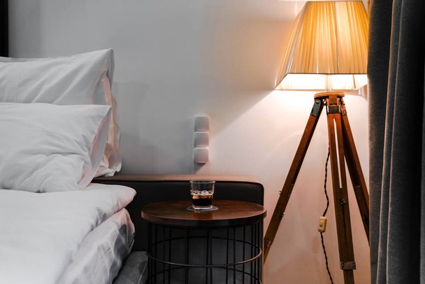 寝室のインテリア。ベッドサイドのテーブルの上にウィスキーを1杯床灯からの柔らかな光が - 写真・画像