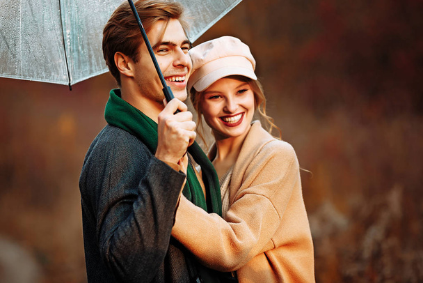 szczęśliwy, para w miłości przytula i uśmiecha się na ulicy, jesień, zielony szalik i czapka, mężczyzna i kobieta na randce, Walentynki. spacer po parku pod parasolem w promieniach zachodzącego słońca - Zdjęcie, obraz
