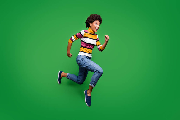 Full body profile side photo of crazy cheerful afro american girl jump run after fall discounts feel rejoice emotions wear bright clothing blue sneakers isolated over green color background - Foto, Imagem