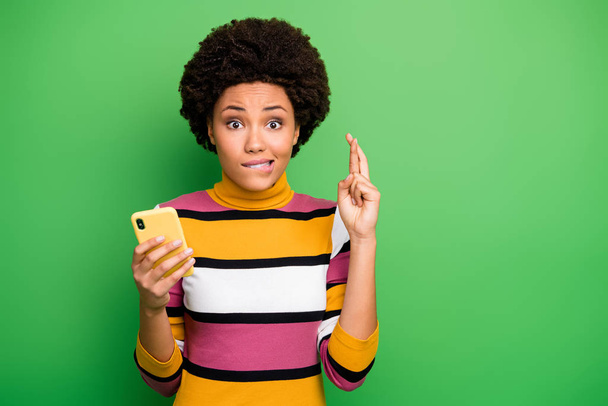 Closeup photo of funny dark skin wavy lady hold telephone fingers crossed wait startup investment results bite lips worried wear casual striped jumper isolated green color background - Foto, afbeelding