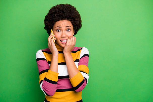 Photo of frightened dark skin wavy lady hold telephone speak best friend terrified listening scary story bite fingers wear casual striped jumper jeans isolated green color background - Foto, Imagen