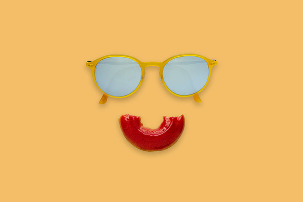 Gele zonnebril en half bijten rode donut sets als glimlach mond op oranje achtergrond voor minimale en zomer concept. - Foto, afbeelding