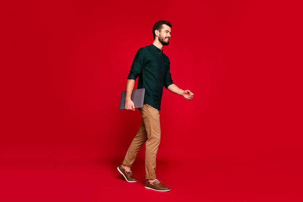 Full length profile side photo of positive charming guy go walk work enterprise hold computer feel he professional expert wear casual style outfit isolated over red color background - Фото, зображення