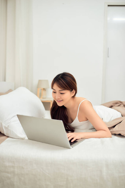 Mosolygó ázsiai fiatal nő használ laptop az ágyban otthon - Fotó, kép