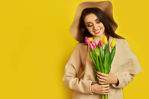 Beautiful girl  dressed in a beautiful coat and hat, with flowers tulips in hands over yellow background. Young woman received flowers as gift and rejoices. Spring girl with flowers. March 8.  - Фото, изображение