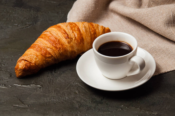 Croissant en kopje koffie en schotel op donkere achtergrond met een grijs linnen servet - Foto, afbeelding