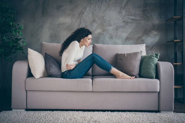 Profile side view of her she attractive depressed unhealthy wavy-haired girl sitting on divan suffering from medical belly problem in modern loft industrial style interior living-room indoors - Photo, image