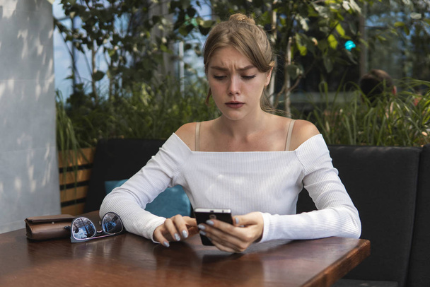 Cheerful blond woman blogger is reading incoming sms message on smartphone connected to free wifi in cafe. Positive young student watching broadcasting online on modern mobile phone. - Фото, изображение
