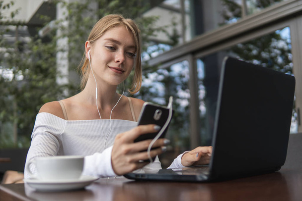 Modern business woman is reading incoming sms message on smartphone or watching broadcasting online on modern mobile phone while sitting in caffe with a laptop. Female student sitting in coffee. - Fotoğraf, Görsel