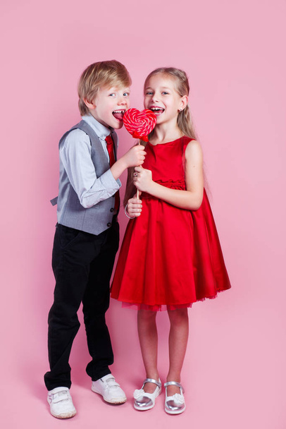 Funny boy and little girl with candy red lollipop in heart shape. Beautiful children eat sweets. Valentines day - Foto, Imagem